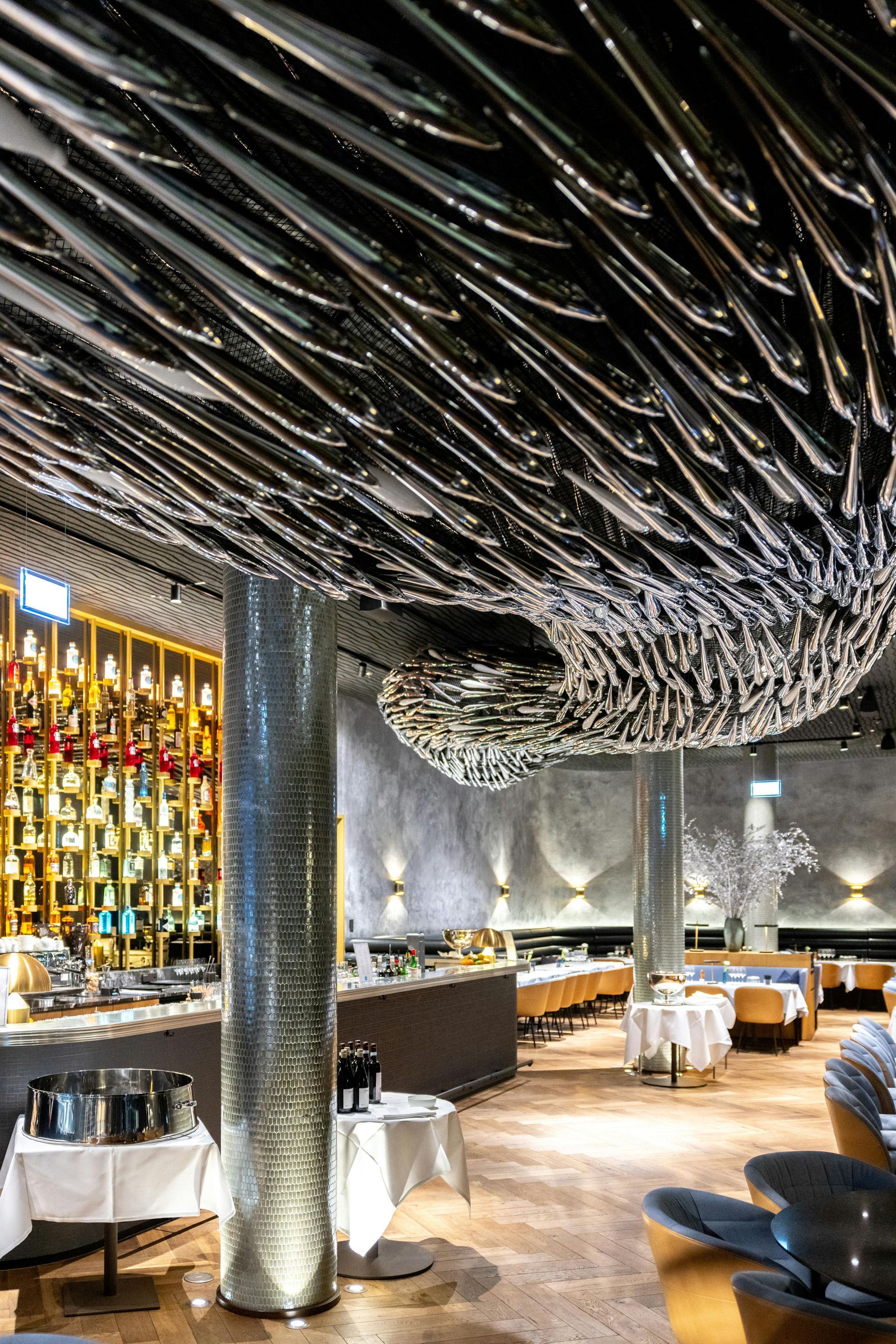 Upscale restaurant interior with a wavy black ceiling, silver column, glowing bar, and white table settings