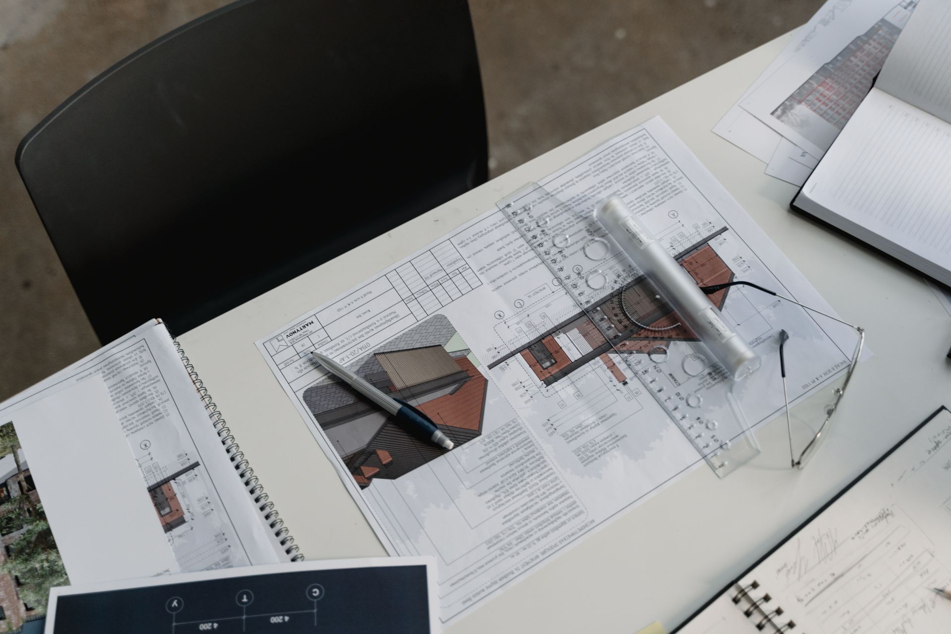 Desk with open notebooks, pens, and papers spread across a workspace near a black chair.