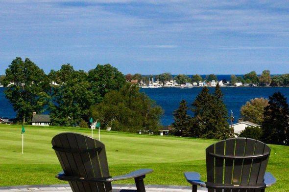 Two Adirondack chairs overlooking a green lawn and blue bay with boats and trees in the distance