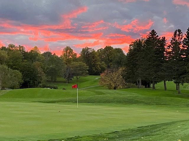 Golf green at sunset with a red flag, trees, and a pink-orange sky