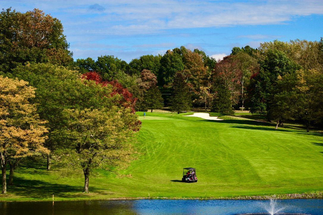 Green golf course bordered by autumn trees, with a water hazard in the foreground.