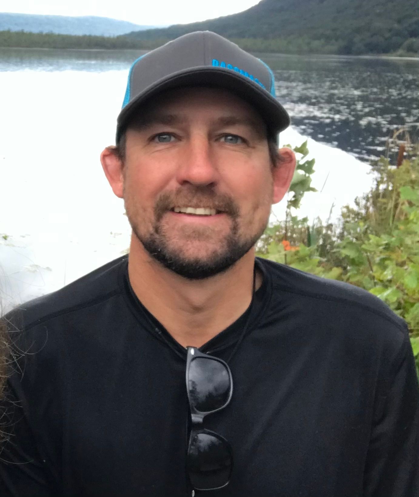 Man wearing a black cap and shirt, smiling by a lakeside with forested hills behind him