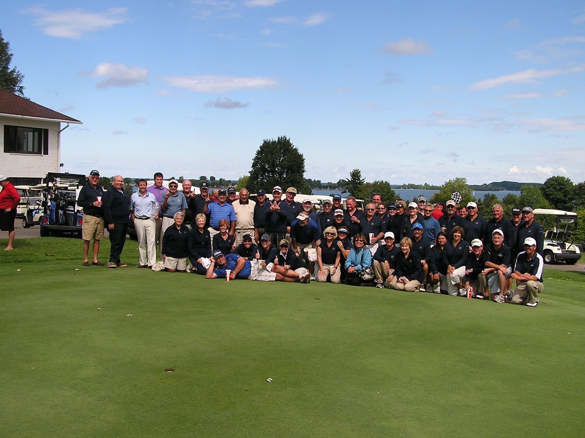 Large group posing on a golf course with a clubhouse and blue sky in the background