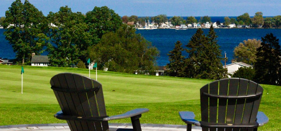 Two black Adirondack chairs overlooking a green lawn and blue lake on a sunny day