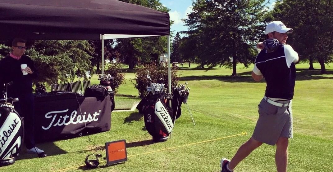 Golfer mid-swing on a grassy tee beside Titleist golf bags under a shaded canopy