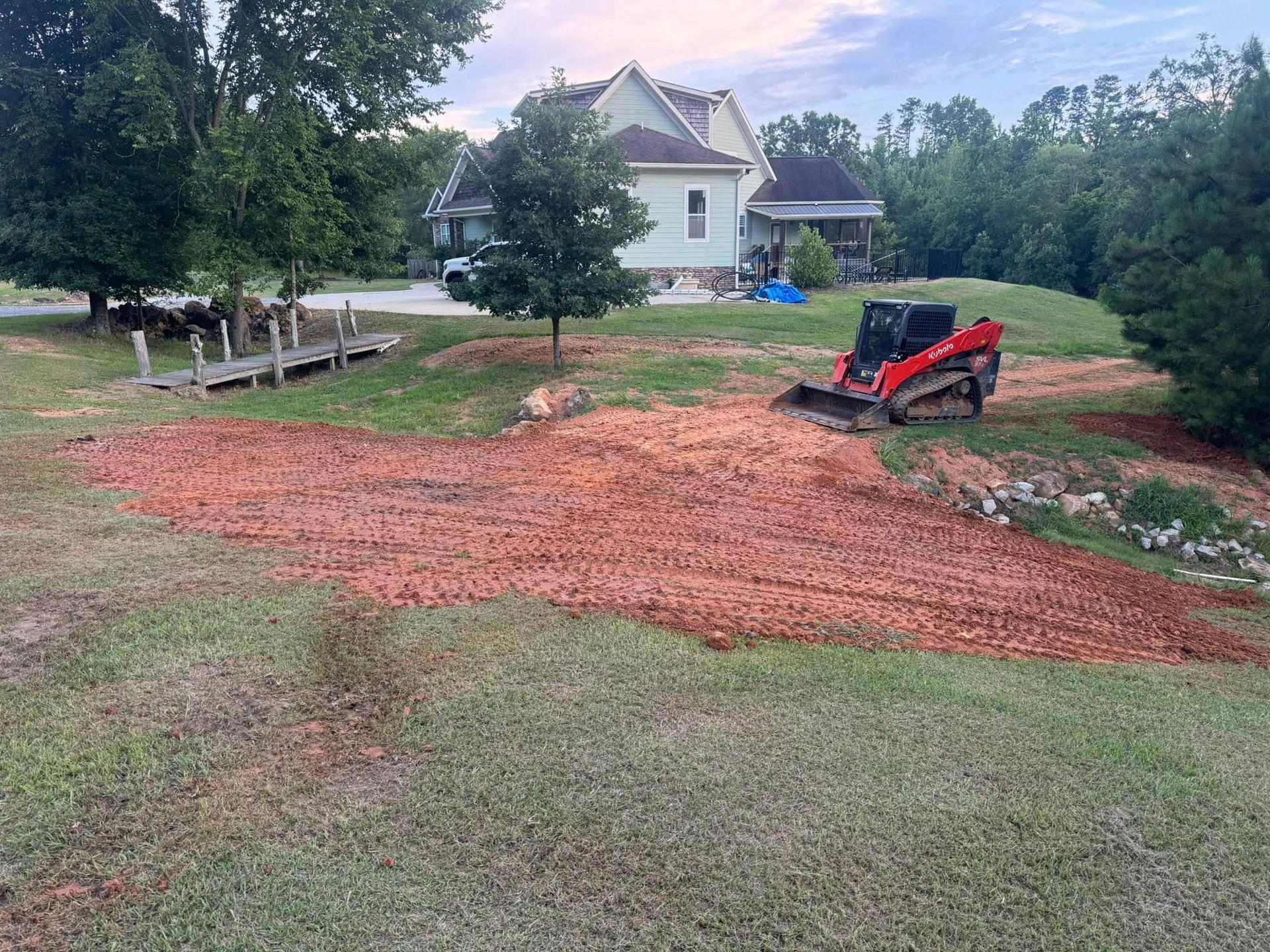Red soil being graded by a red skid steer on a grassy hill with a house and trees in the background.