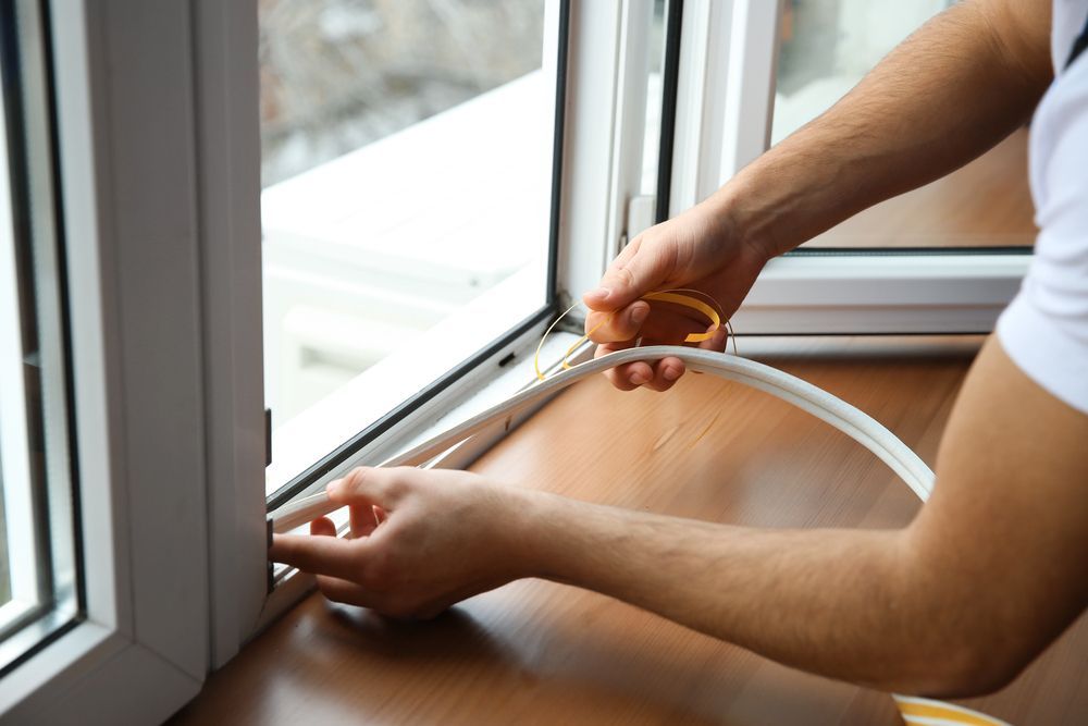 Persona che installa guarnizioni attorno al telaio di una finestra. Profilo bianco, striscia di schiuma beige, superficie in legno.