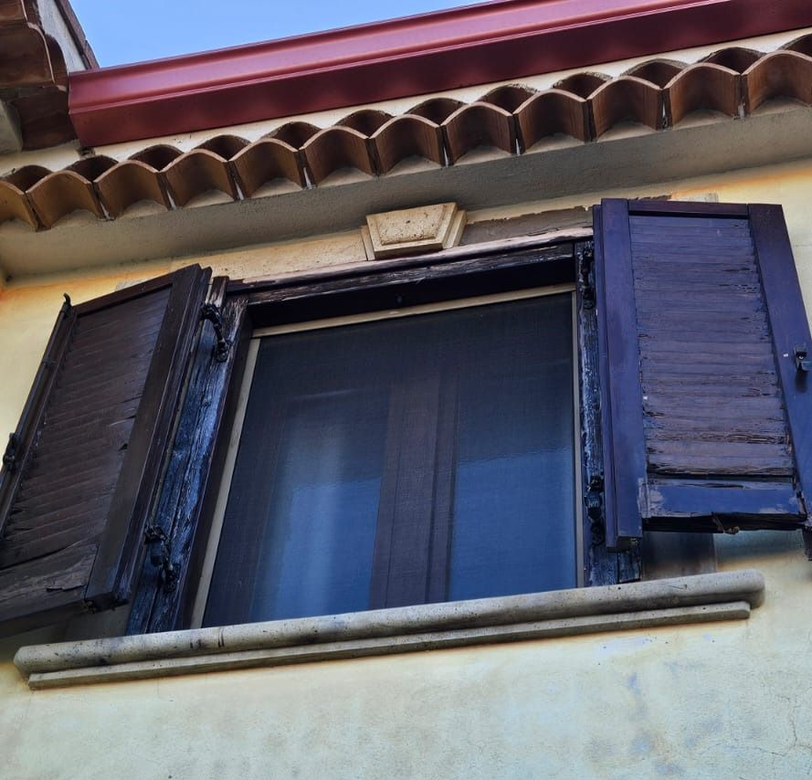 Finestra con persiane di legno scuro aperte su un edificio beige con tetto di tegole rosse.