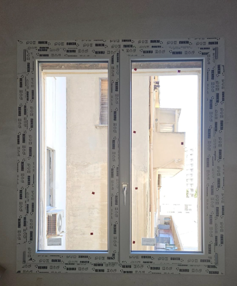 Finestra bianca con doppi vetri; vista sugli edifici e sullo spazio esterno; montata su parete bianca.