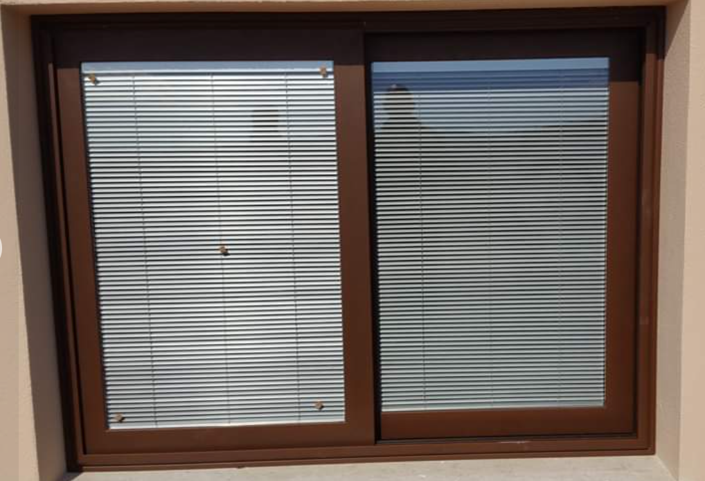 Finestra scorrevole con cornice marrone e persiane orizzontali chiuse; luce naturale all'esterno.