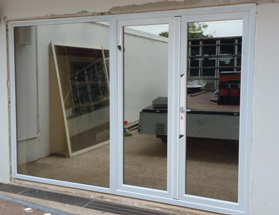 Porte in vetro con cornice bianca che riflettono una stanza con oggetti e utensili, inserite nell'apertura di un edificio.