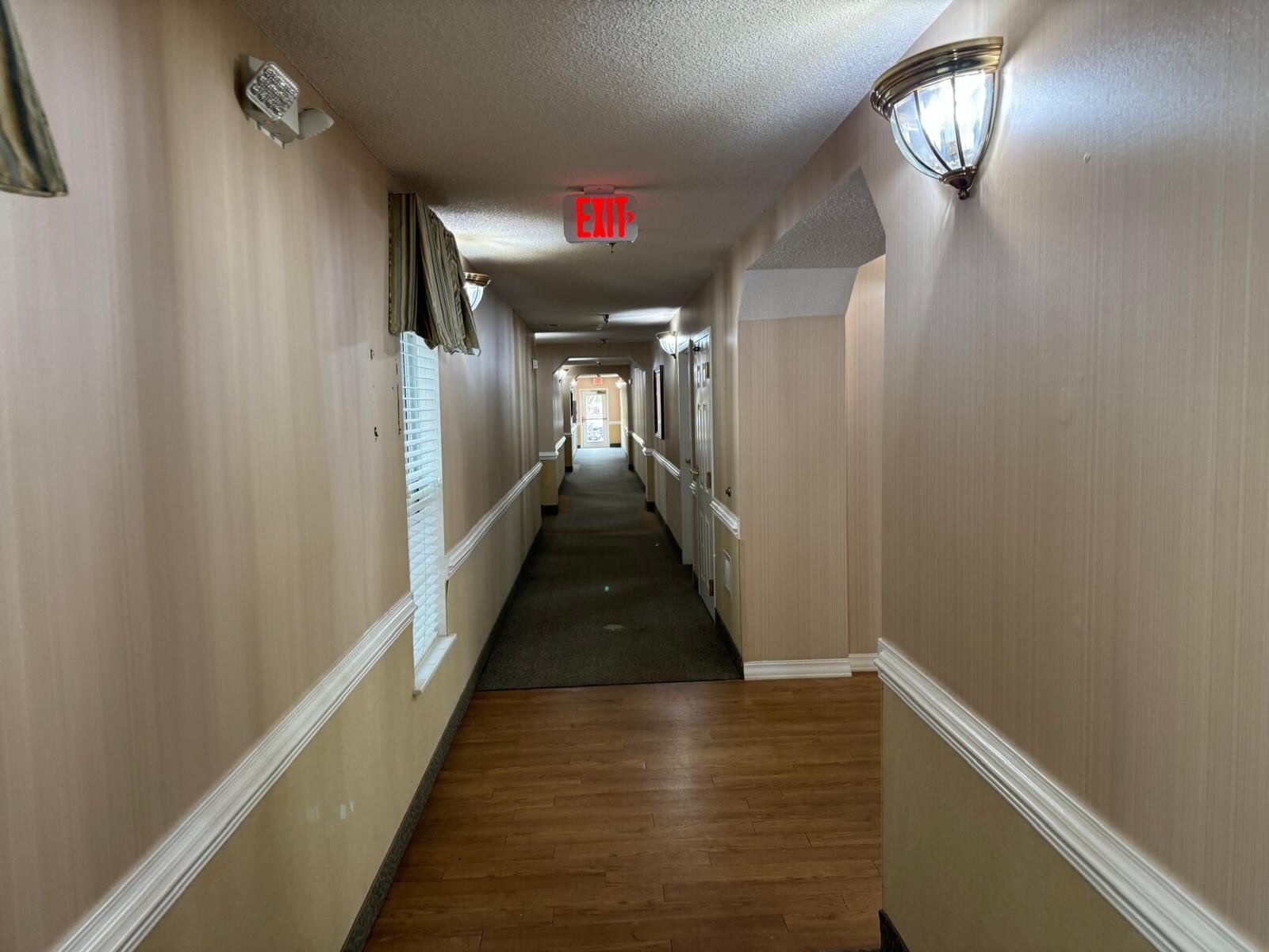 Dim hotel hallway with beige walls, wood floor, wall sconces, and red EXIT sign at the end.