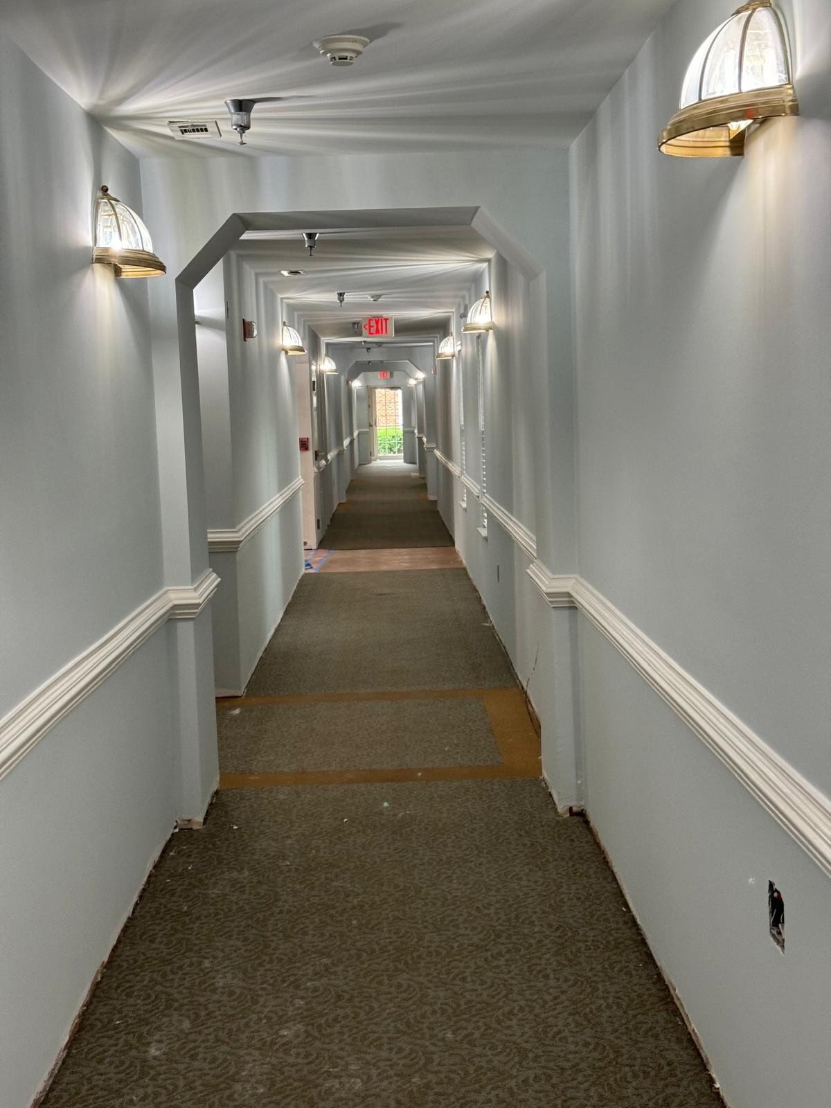 Long, narrow hallway with gray walls, wall lamps, and carpeted floor leading to a distant exit.