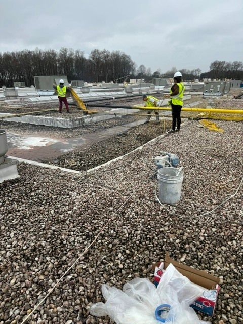 Two workers in yellow vests on a gravel construction site, handling yellow cables near concrete slabs.
