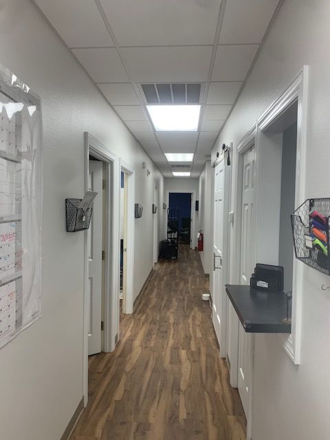 Narrow office hallway with white walls, wood floor, ceiling lights, and open doorways on both sides.