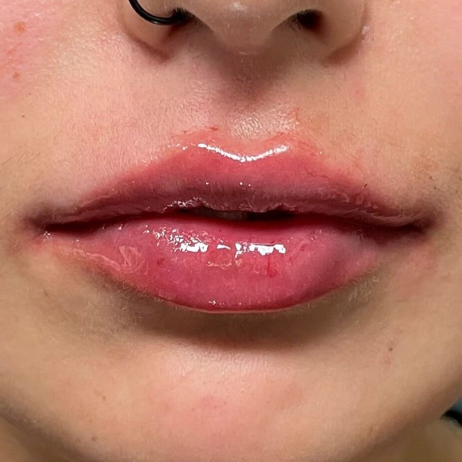 Close-up of a person's lips. Shiny, plump, reddish-pink lips with a slight dip in the upper lip. A nose ring is visible.