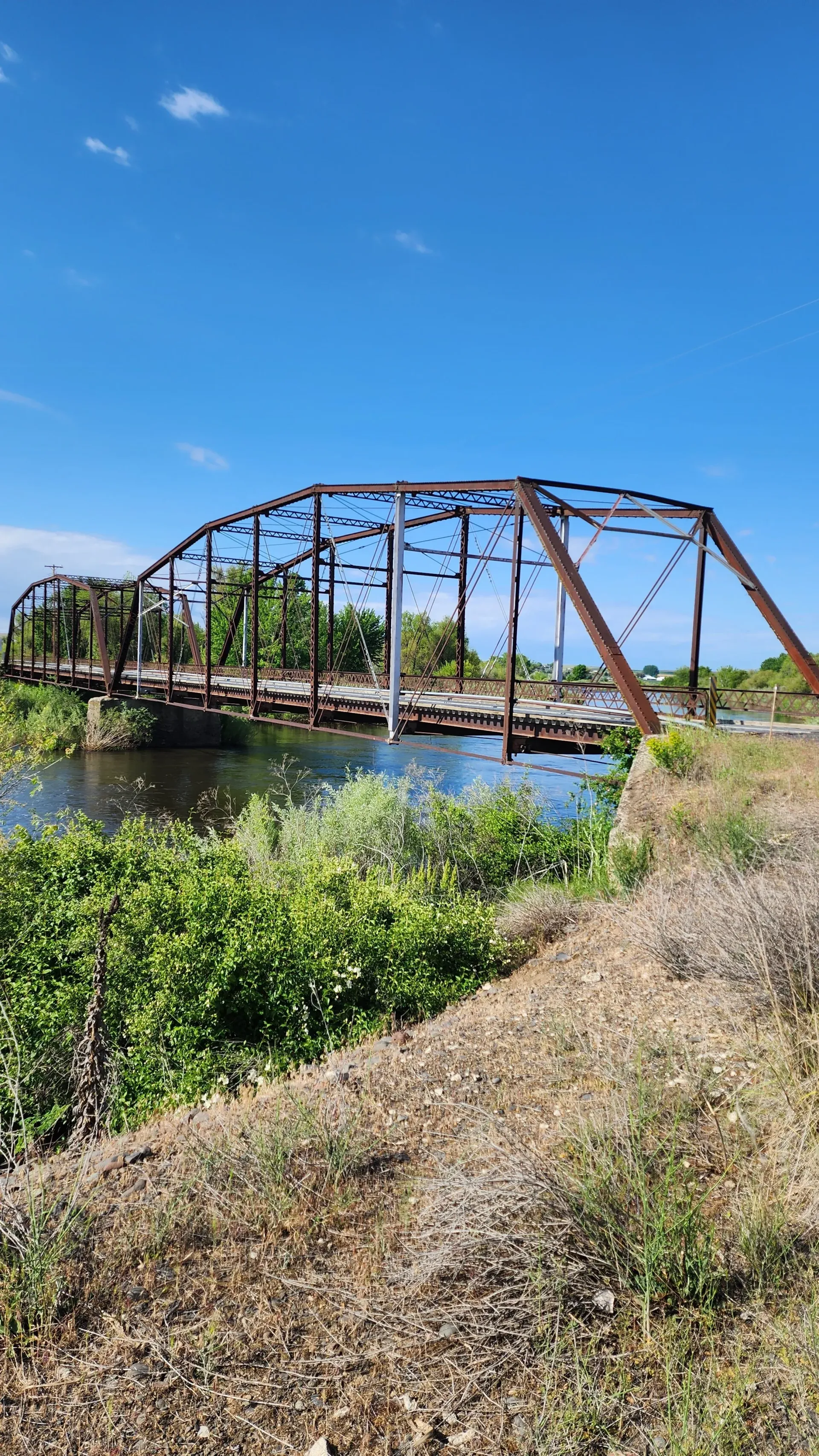Rusty truss bridge over a calm river, viewed from a grassy bank under a blue sky.