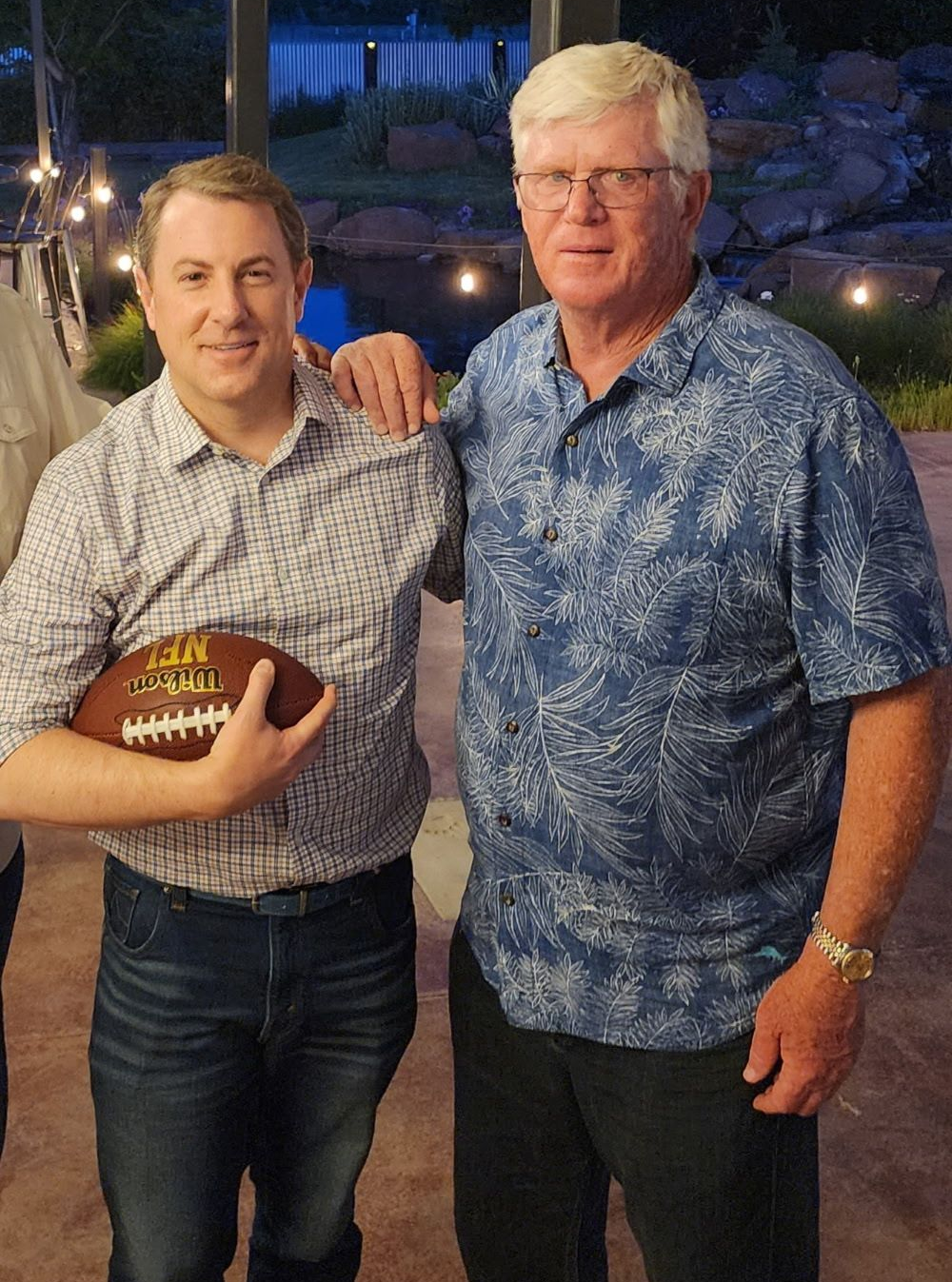 Two men standing together indoors, one holding a football, both smiling at a nighttime event.