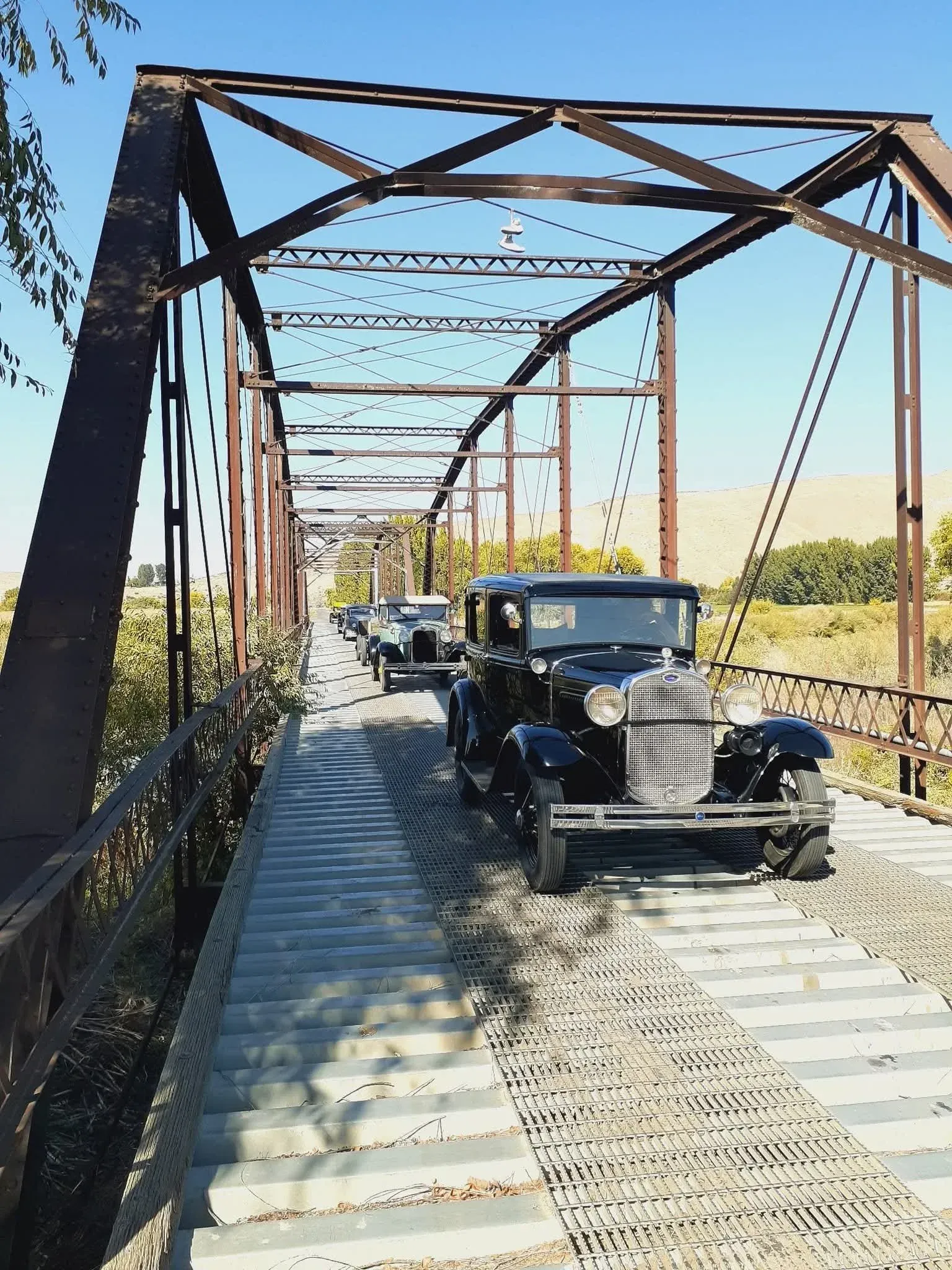 Vintage cars crossing a rusty metal truss bridge on a sunny day