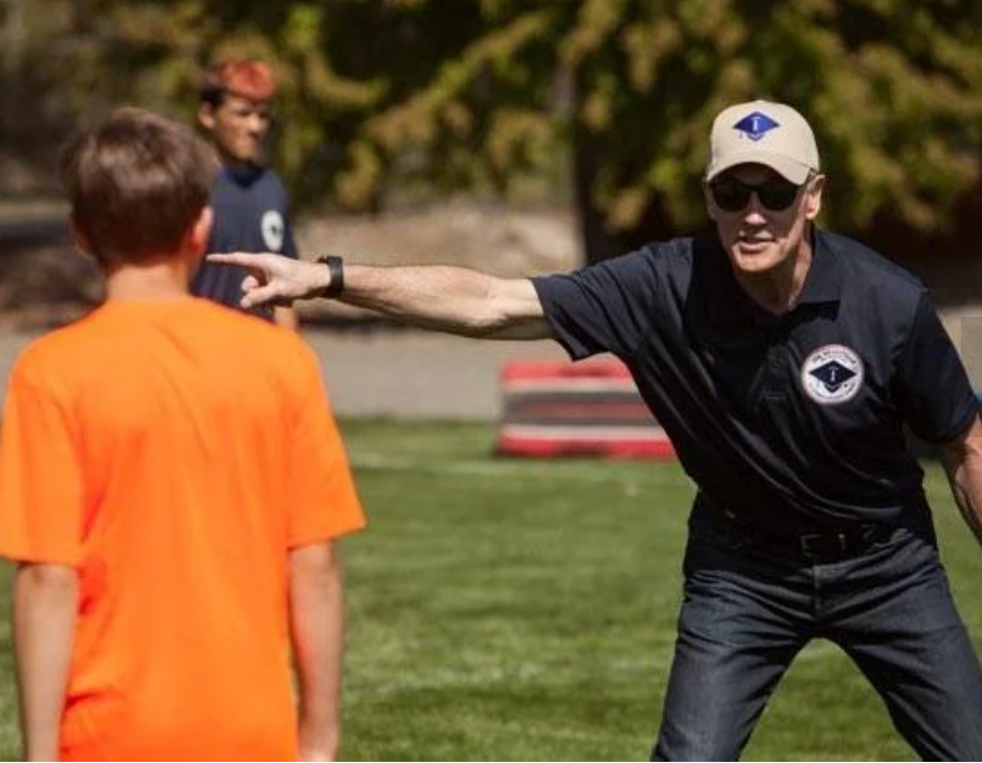 Coach in black shirt and cap points at a player in an orange shirt on a grassy field