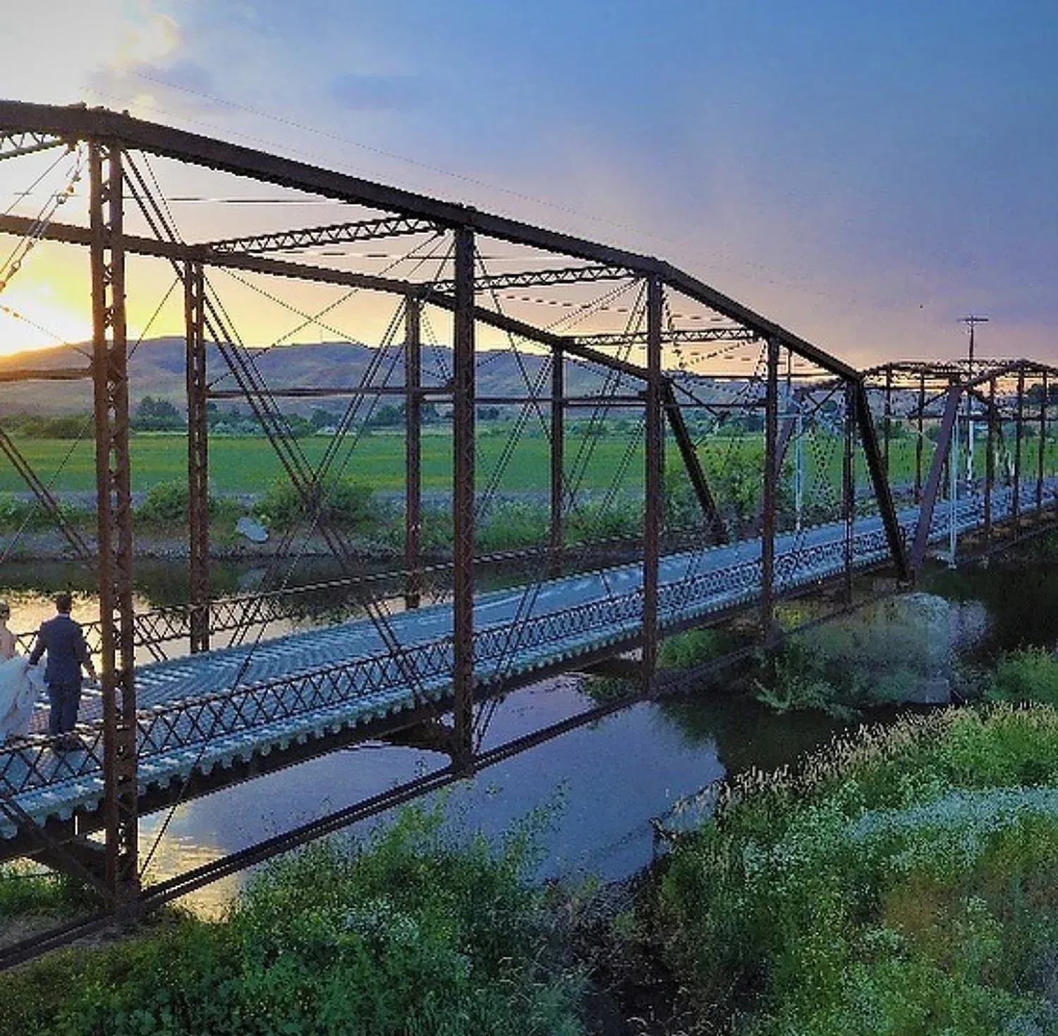 Narrow metal truss bridge over a canal at sunset, with green fields and distant hills.
