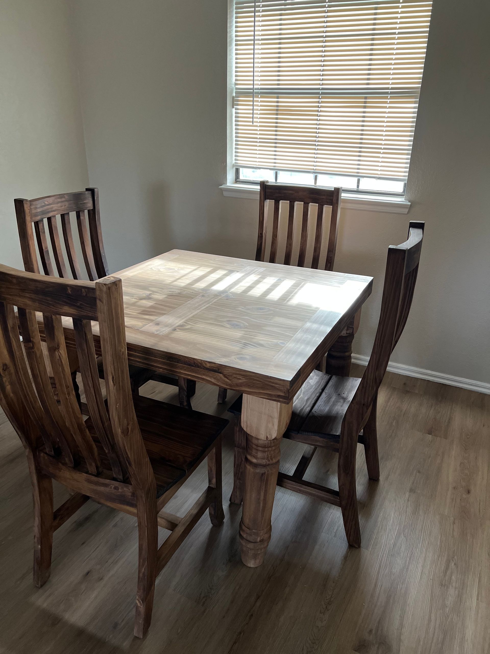 A square wooden dining table with four matching chairs sits by a window with blinds in a room.