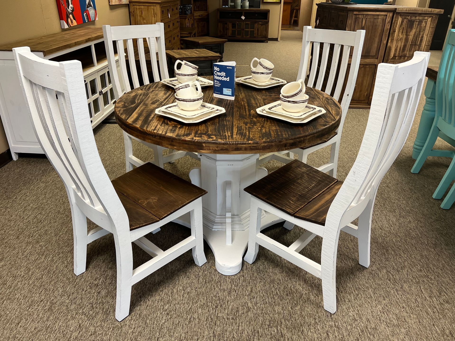 A round dining table with four white chairs and teacups, set in a store with wood furniture.