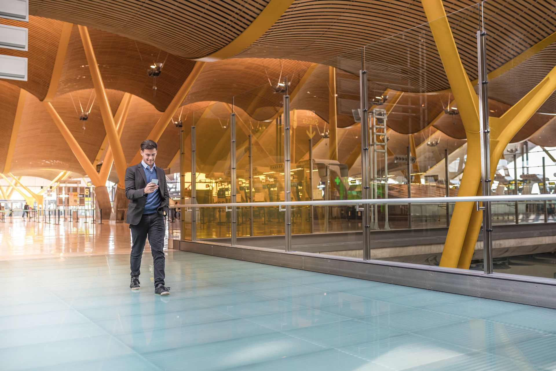 Man in suit walks through airport, looking at phone. Wooden ceiling and yellow support columns.