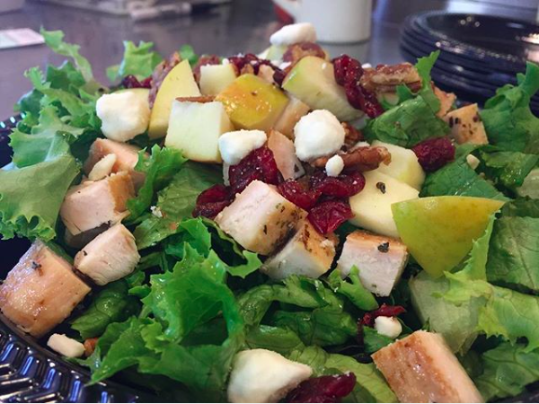 Fresh salad with greens, grilled chicken, croutons, cranberries, and yellow fruit in a black plastic bowl