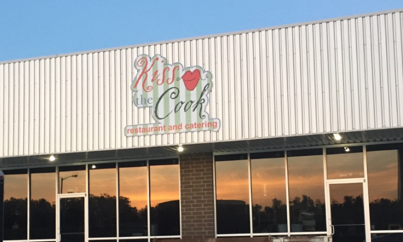 Storefront sign reading “Kiss My Cook” on a building at sunset.