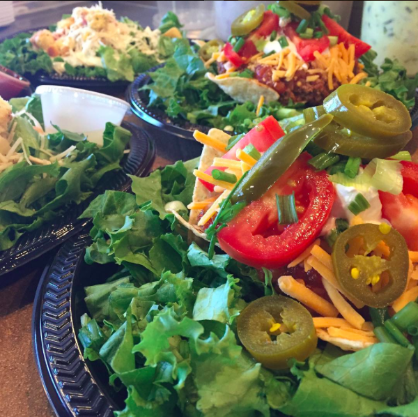 Fresh salad bar with leafy greens, tomato slices, jalapeños, shredded cheese, and other toppings in black bowls