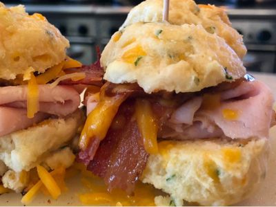 Hands holding a bacon, cheese, and biscuit breakfast sandwich with melted cheddar.