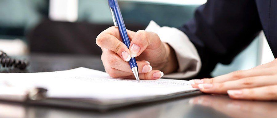 Manos firmando un documento con un bolígrafo azul en un escritorio.
