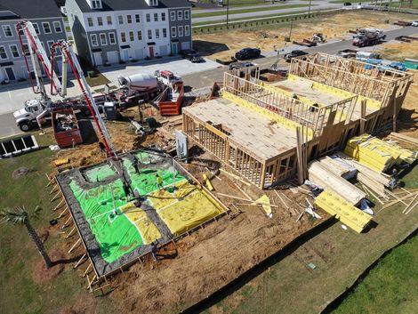 Aerial view of a house under construction with framing, concrete slabs, and a crane on muddy ground