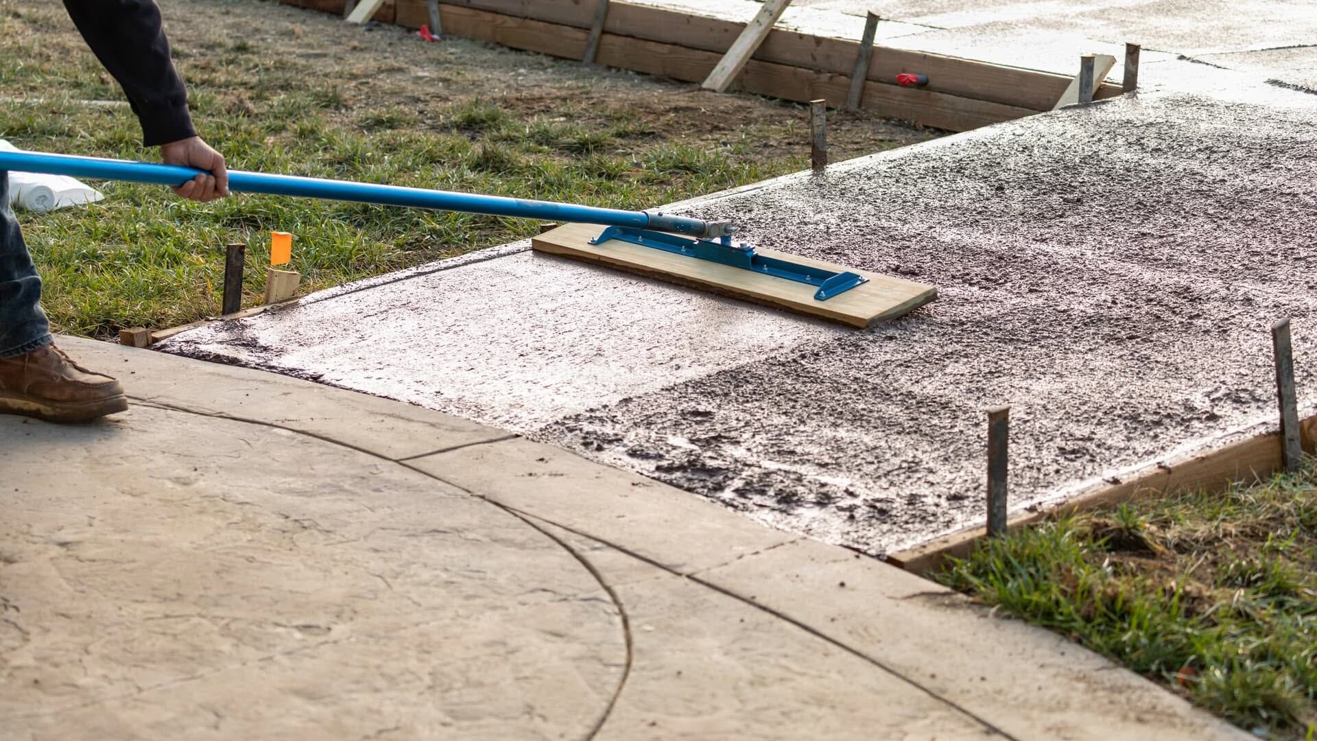 Worker smoothing wet concrete on a sidewalk beside a grassy area and curb