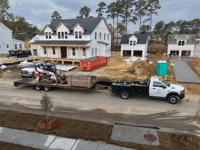 Construction site with white houses, trucks, and an orange dumpster beside muddy lots