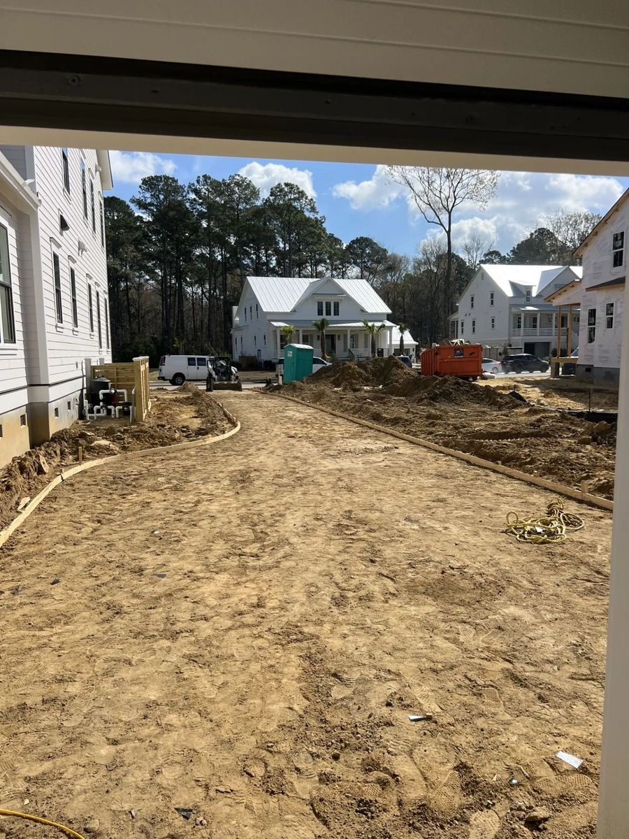 Construction site with dirt roadway between white houses, trees, and equipment under a partly cloudy sky