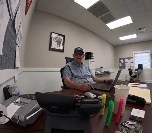 Man sitting at an office desk with laptop, phone, and office supplies in a bright workspace