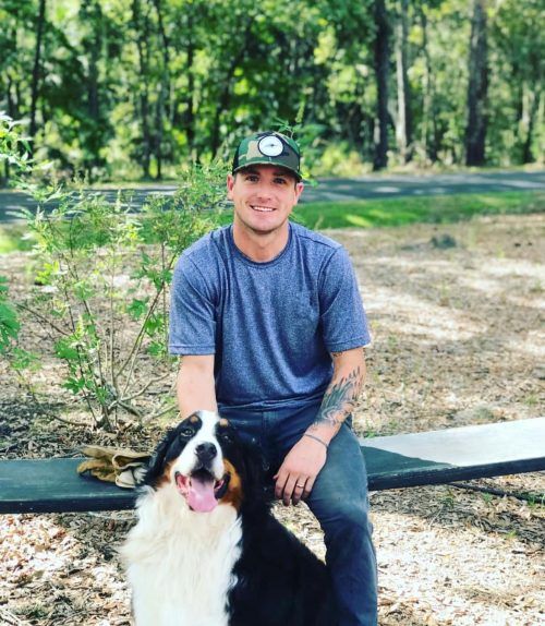 Man in blue shirt sitting outdoors with a black-and-white dog on a bench, trees in the background