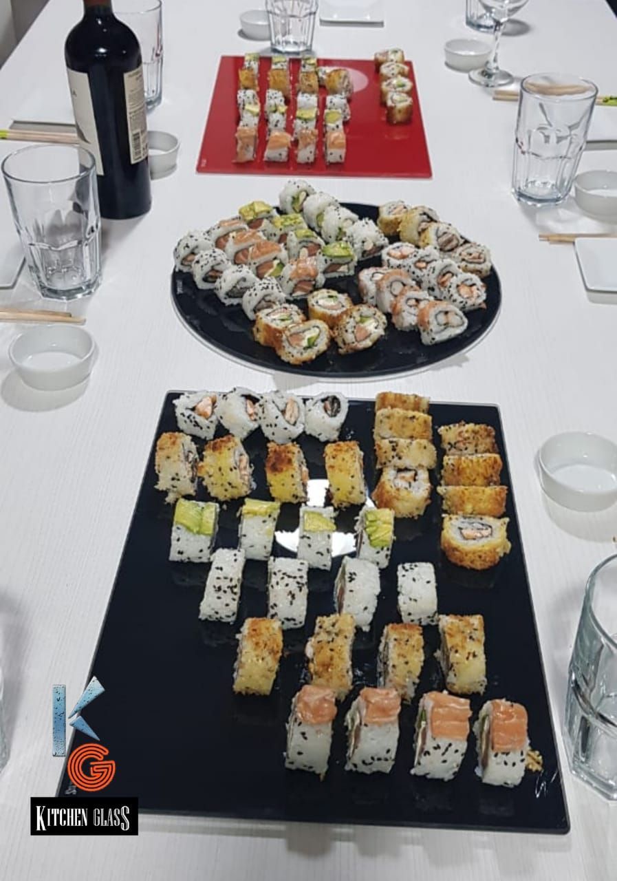 Plato de sushi sobre una mesa, varios rollos, una botella de vino, vasos, palillos y cuencos para mojar.