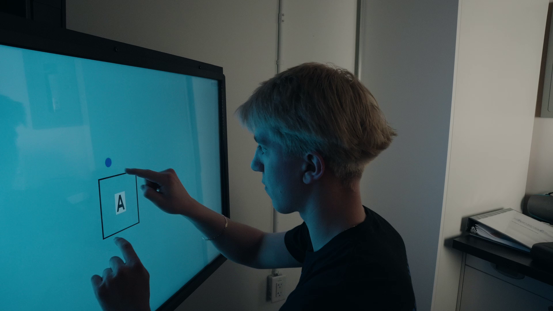 A person interacts with a blue digital screen, using both hands to manipulate a box containing the letter A.