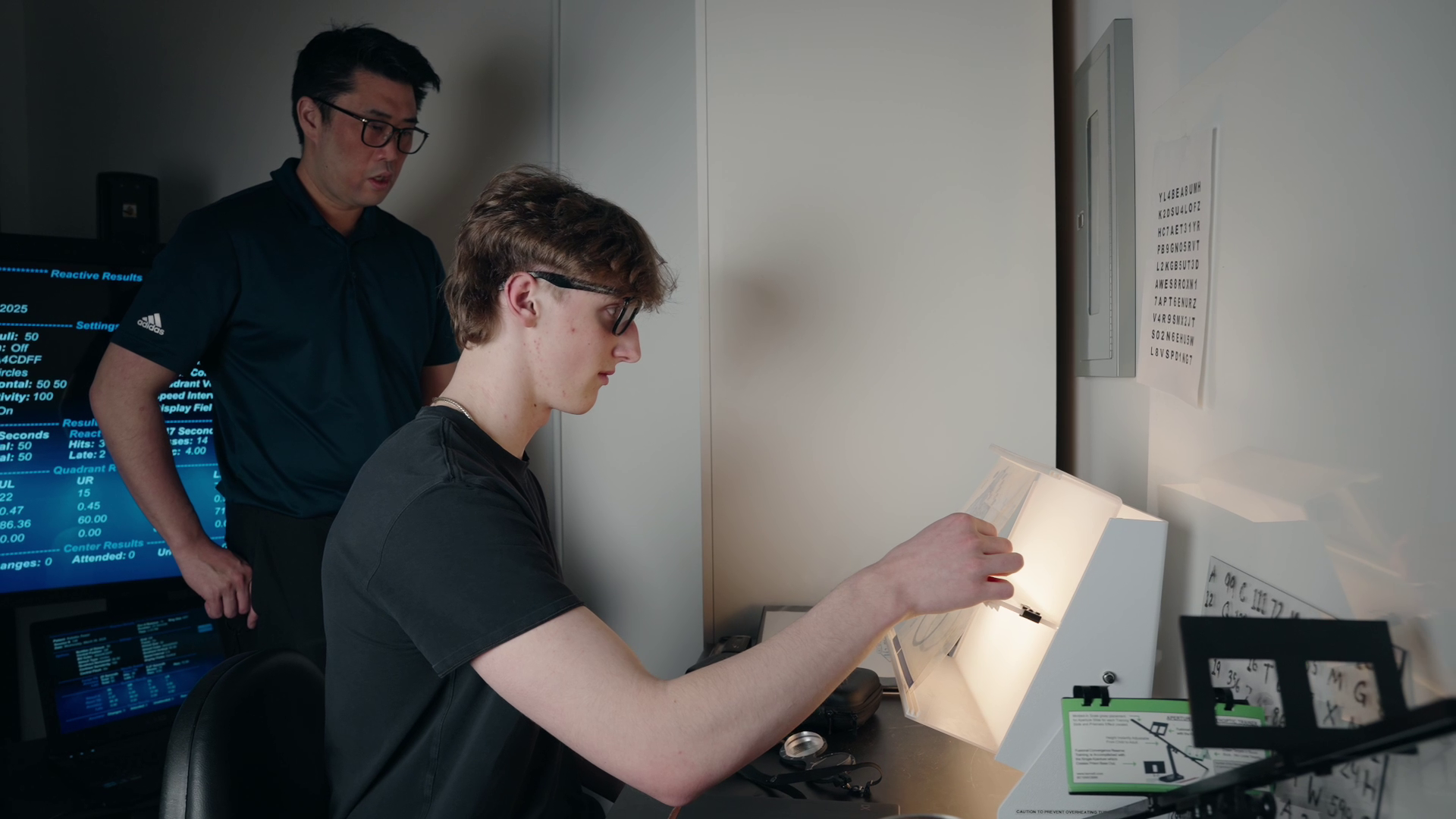 An individual sits at a desk using an illuminated eye-testing device while another person observes from behind.
