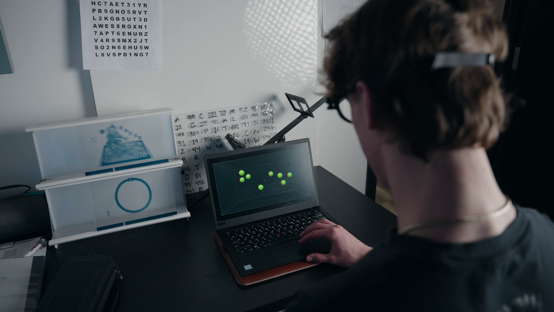 A person wearing a head-mounted device works at a desk, looking at a laptop screen displaying a pattern of green dots.