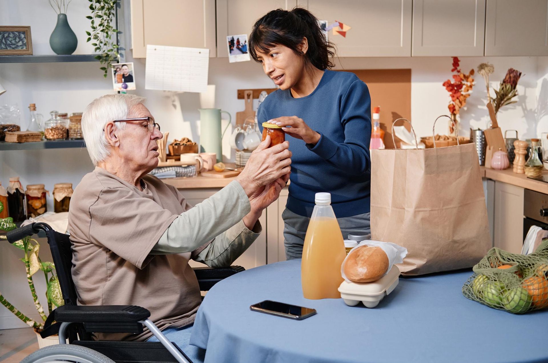 Woman helping an elderly person in a wheelchair with groceries in a kitchen.
