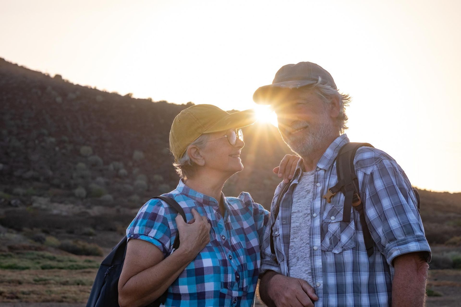 Couple hikers smiling at sunset, wearing hats and backpacks, outdoors.