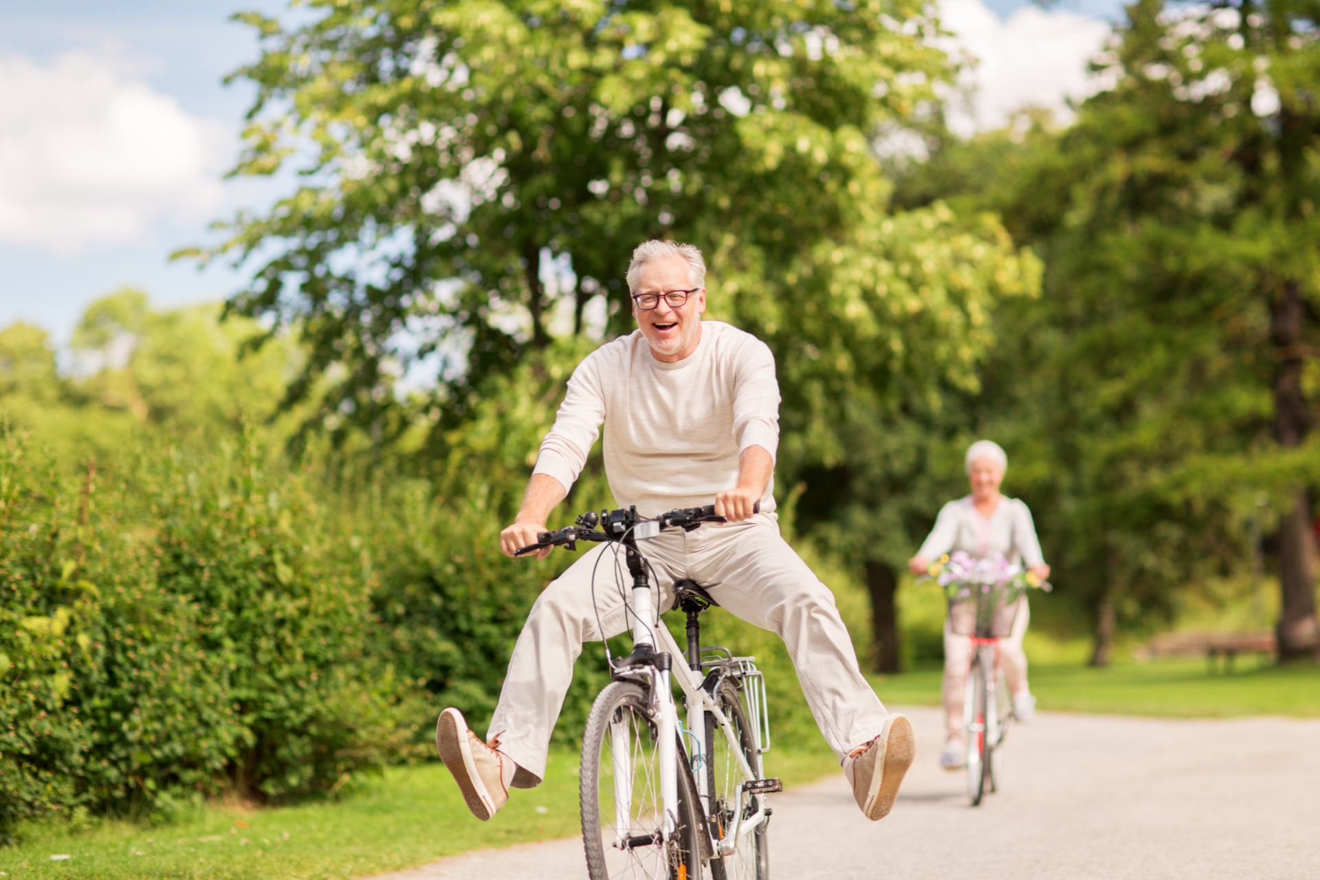 Smiling person riding a bike with legs outstretched, followed by another person biking on a paved path in a park.