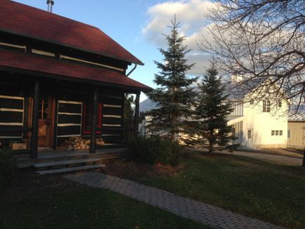 A log cabin with a porch and a white house in the background