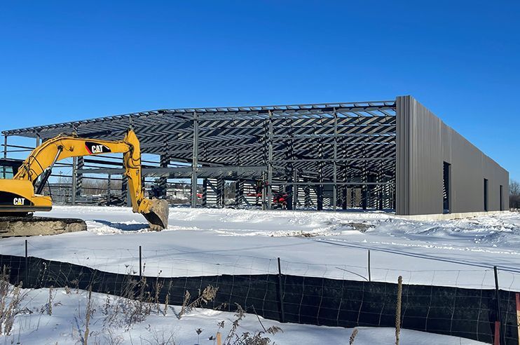 A yellow excavator is digging in the snow in front of a building under construction.