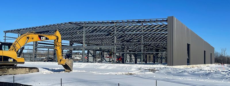A large building is being built in the snow with a yellow excavator in front of it.