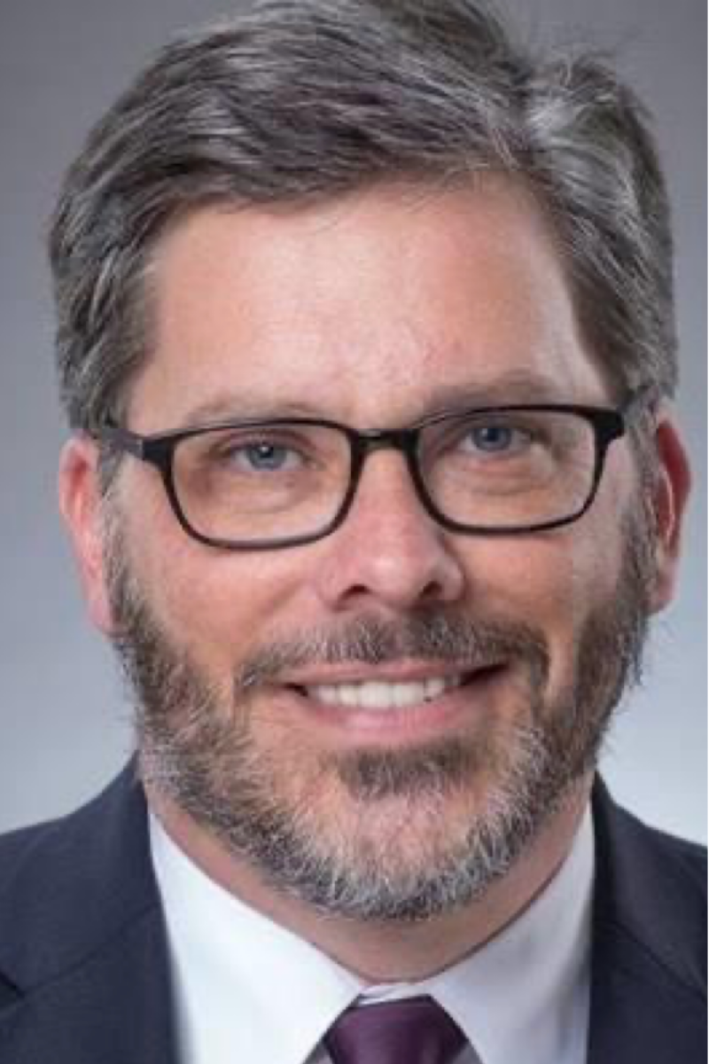 A man with a beard and glasses is wearing a suit and tie and smiling.